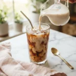 Summer moon moon milk recipe swirling into a glass of iced coffee on a marble counter